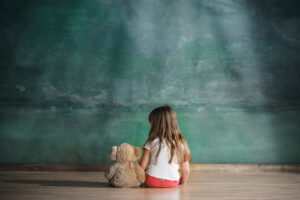 Image of a girl with a teddy bear sitting in front of a chalk board. Showing how a kid can feel isolated without children's therapy in Maryland. We use play therapy with toys such as the bear as part of our child therapy in the Washington DC area and Baltimore, MD.